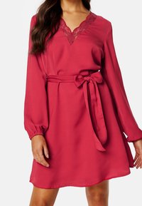 Robe rouge à manches longues, décolleté en V en dentelle, et une ceinture assortie nouée à la taille. Tissu lisse avec une coupe décontractée et un ourlet évasé.