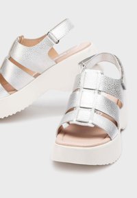 Sandalias metálicas plateadas con una suela gruesa blanca, con puntera abierta, correa ajustable en el tobillo y múltiples correas horizontales. Textura suave.