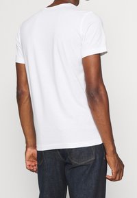 Camiseta blanca de algodón con cuello redondo, mangas cortas y corte relajado, vista desde atrás, combinada con vaqueros de denim azul oscuro.
