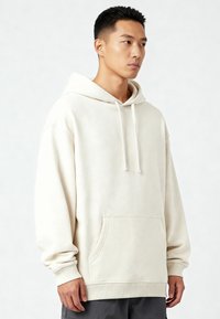 Krém színű kapucnis pulóver puha anyagból, elülső zsebbel, bordázott mandzsettákkal és laza fitettel. Finom textúrával és zsinóros kapucnival rendelkezik.