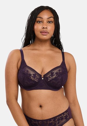 Mujer curvilínea con el cabello trenzado que lleva un sujetador de encaje morado oscuro y ropa interior de encaje a juego, posando frente a un fondo blanco liso.