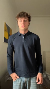 Mlad moški s kodrastimi lasmi, oblečen v mornarsko modro polo majico z dolgimi rokavi in svetlo modre kavbojke, stoji v notranjosti s rokami v žepih.