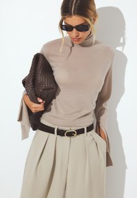 Beige Rollkragenpullover mit weit ausgestellten Ärmeln, kombiniert mit hellbeigen, gefaltetem Hosen und einer gewebten braunen Clutch. Schwarzer Gürtel mit goldenen Akzenten.