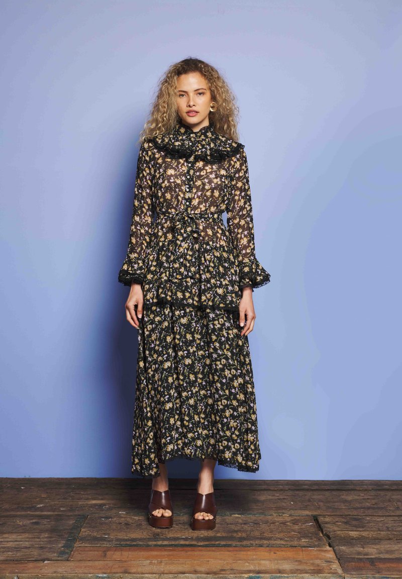 Göndör hajú nő, hosszú, áttetsző fekete virágmintás ruhában és barna platform szandálban áll egy fából készült padlón, kék fal előtt.