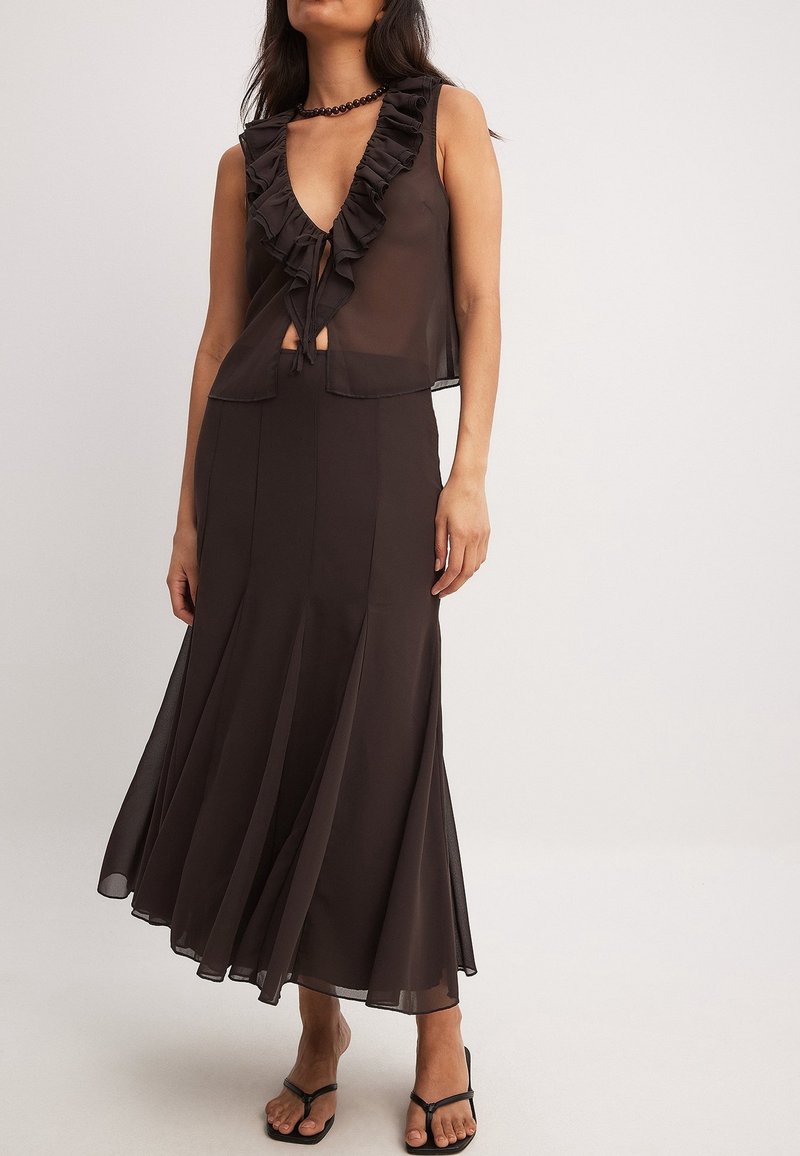 Femme portant un haut noir transparent sans manches avec des volants et une longue jupe fluide assortie avec des sandales noires sur un fond uni.