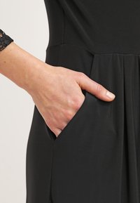 Main partiellement insérée dans la poche latérale d'un vêtement noir avec des détails plissés près de la taille.