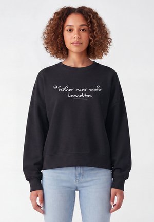 Frau mit lockigem braunem Haar trägt einen schwarzen Pullover mit deutschem Text und hellblauen Jeans, stehend vor einem schlichten weißen Hintergrund.
