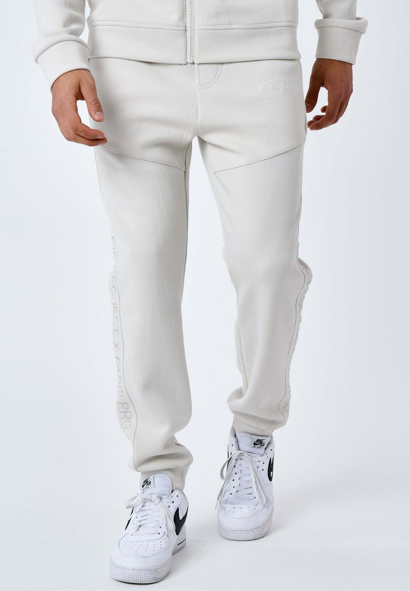 Pantalon de survêtement gris clair avec une texture lisse, doté de poches latérales inclinées et d'une marque discrète brodée sur le côté. Associé à des baskets blanches.