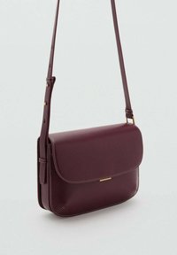 Borsa a tracolla in pelle bordeaux scuro con tracolla regolabile, patella curva e delicato gancio dorato su sfondo grigio chiaro.