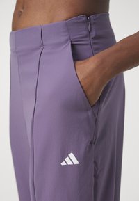 Fioletowe spodnie sportowe wykonane z lekkiego, gładkiego materiału, z boczną kieszenią i białym logo Adidas po lewej stronie.