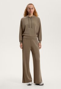 Rjava kapuca s kapuco z vrvico, sproščen kroj in široke hlače v ujemajoči barvi, izdelane iz mehkega materiala; obuta z lahkimi športnimi copati.