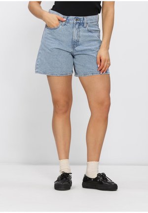 Mujer con pantalones cortos de mezclilla azul claro, calcetas color crema y zapatos negros con cordones, de pie con una mano en el bolsillo contra un fondo blanco.