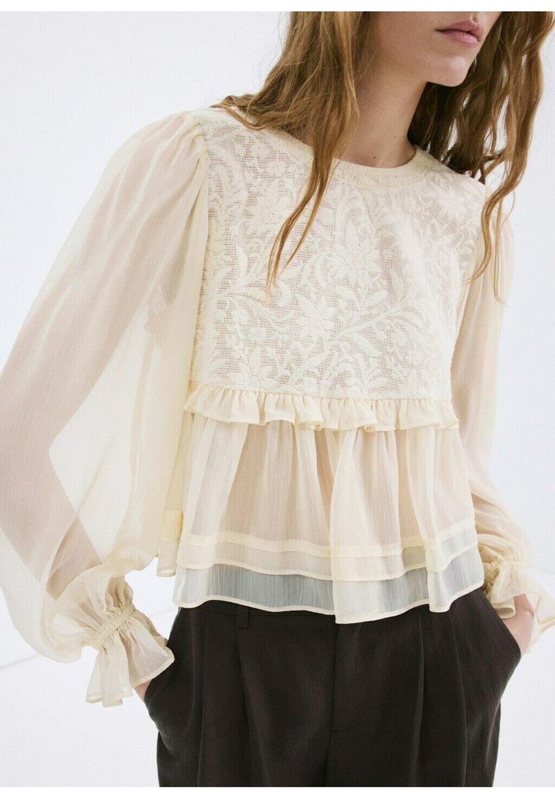 Femme portant un chemisier à manches longues en dentelle crème transparente avec des volants, rentré dans un pantalon noir taille haute, mains dans les poches.