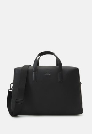 Sac de sport noir avec deux poignées supérieures, une bandoulière détachable, texture lisse et logo Calvin Klein. Forme rectangulaire compacte.