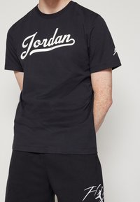 Osoba oblečená v černém tričku Jordan s bílým logem a černých šortkách s bílou grafikou na levé noze, stojí s rukama za zády.