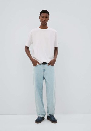 Giovane uomo in piedi con le mani nelle tasche dei jeans azzurri, indossa una maglietta bianca e sneakers scure su uno sfondo chiaro e uniforme.
