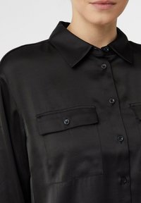 Close-up of a person wearing a black button-up shirt with two chest pockets and a pointed collar.