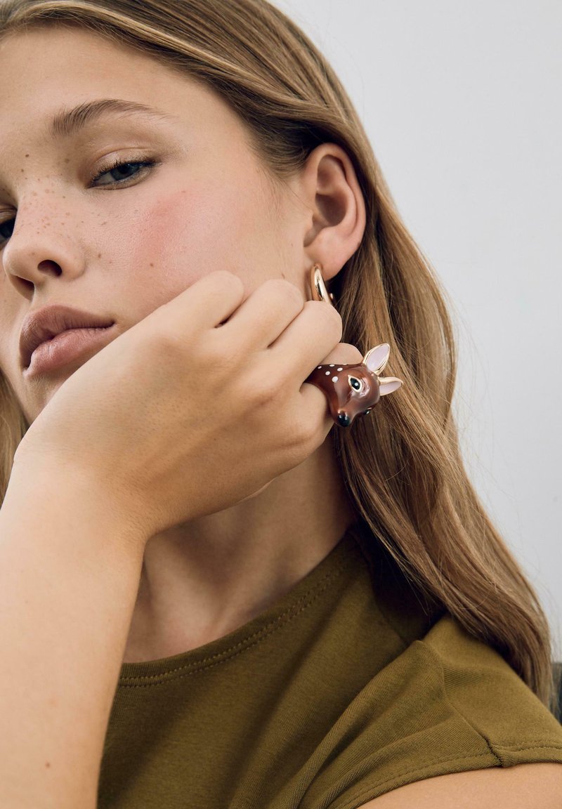 Anello a forma di cervo marrone con dettagli dipinti, indossato sull'indice. Il modello ha capelli castani chiari e indossa una camicia verde oliva.