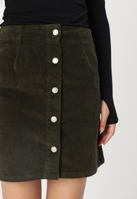 Falda de pana color verde oliva con corte recto, que presenta cinco botones plateados en la parte delantera y un dobladillo que se sitúa por encima de la rodilla.