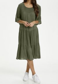 Mujer que lleva un vestido de raíces verdes, de largo a la rodilla, con mangas tres cuartos y zapatillas blancas, de pie con las manos entrelazadas.