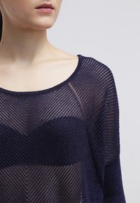 Námorně modrý pletený top s texturovaným, průhledným designem a chevron vzorem. Široký výstřih a dlouhé rukávy. Viditelné jemně odrazující nitě.