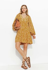 Robe florale jaune avec de longues manches bouffantes et un décolleté en V. Associée à un sac brun clair et des sandales à lanières, présentant un motif léopard.