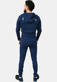 Chándal azul marino hecho de tela suave, con capucha y diseño ajustado. El texto en la parte trasera dice "HOODIE" en amarillo. Zapatillas negras.