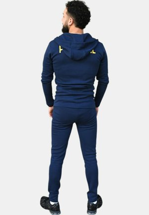 Chándal azul marino hecho de tela suave, con capucha y diseño ajustado. El texto en la parte trasera dice "HOODIE" en amarillo. Zapatillas negras.