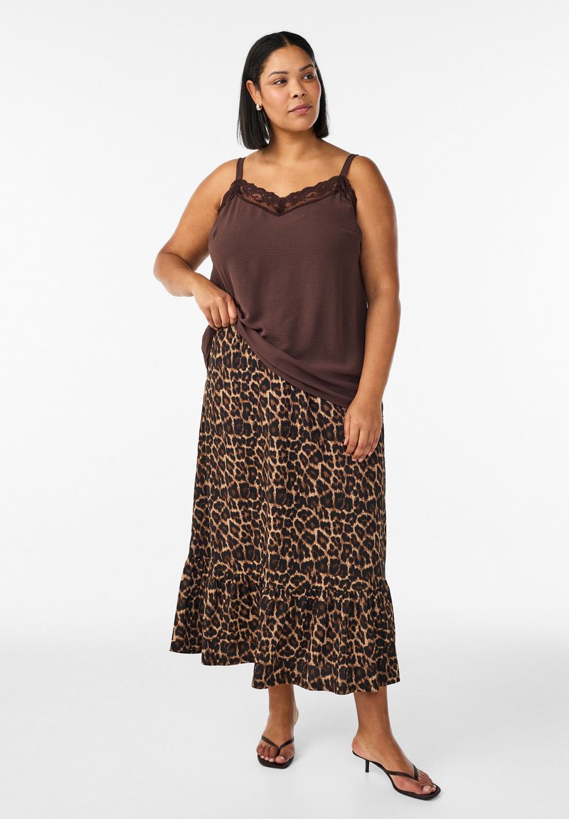 Mujer con cabello hasta los hombros lleva una camiseta sin mangas marrón con encaje, falda de estampado de leopardo y sandalias negras de tacón, de pie con una postura relajada.