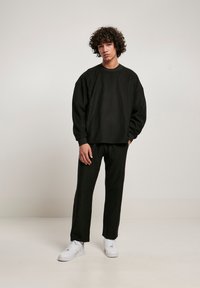 Sort ribbet sweatshirt med lange ærmer og matchende bukser, med en løs pasform og rund hals, parret med hvide sneakers på en ensfarvet baggrund.