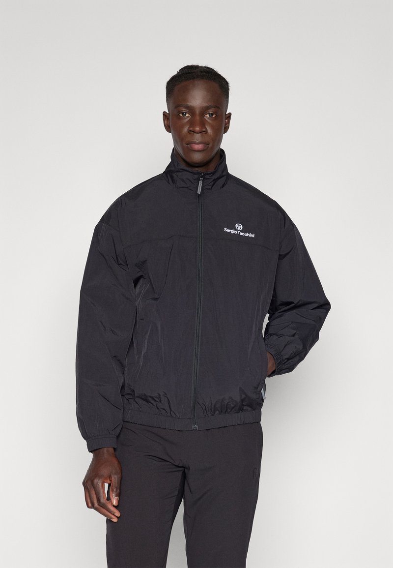Svart zip-up jakke med høy krage, elastiske mansjetter og hem. Har et hvitt logo på brystet. Laget av lett stoff.
