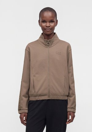 Personne portant une veste marron clair à fermeture éclair avec un col montant et un pantalon noir, debout devant un fond uni clair.