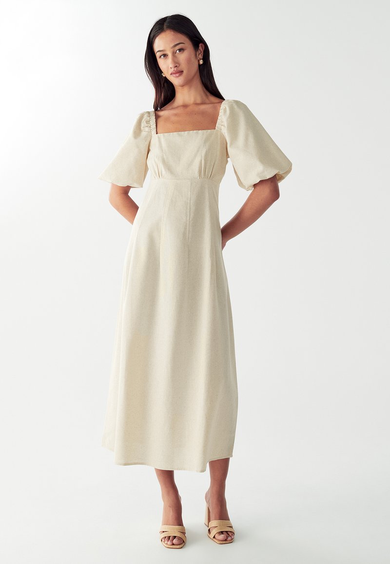 Haljina od mješavine lana krem boje s kvadratnim dekolteom, napučenim rukavima i prilagođenim korzetom. Pada do sredine lista s mekom teksturom.
