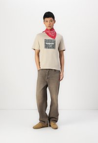 T-shirt beige avec un patch graphique gris, jean ample marron et baskets beige. Un bandana rouge autour du cou ajoute un détail contrastant.