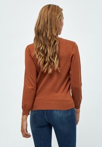 Brauner Strickpullover mit langen Ärmeln, gerippten Bündchen und Saum, körperbetonter Schnitt, über blauen Jeans getragen. Rückansicht, glatte Textur.