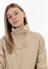 Beige voděodolná bunda s vysokým límcem, vybavená stříbrnými cvoky a hladkou, lehkou texturou. Jednoduchý design bez vzorů.