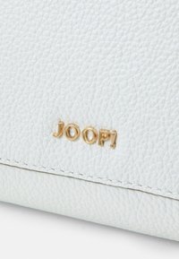 Balta odinė piniginė su tekstūruotu paviršiumi ir auksiniu „JOOP!“ logotipu iškilminiu ant priekio. Siūlės detalizavimas aplink kraštus.