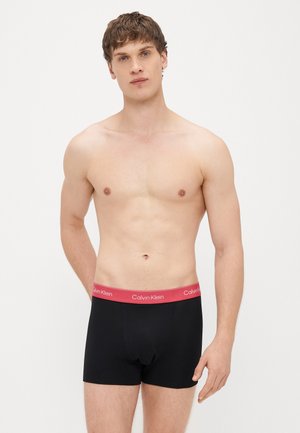 Boxers de algodão pretos com uma cintura elástica vermelha com o logótipo "Calvin Klein", concebidos para um ajuste justo. O modelo está em pé com uma expressão neutra.