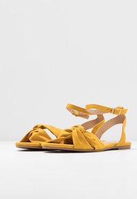 Keltaiset suedesandaalit, joissa on solmittu etuosan muotoilu ja nilkkahihna. Litteä pohja, avokärkinen ja sileä pintakuvio. Yksinkertainen ja moderni muoto.