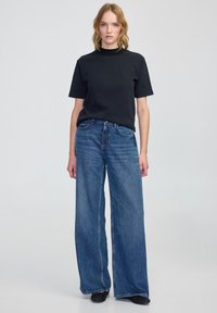 Černé tričko s krátkým rukávem v kombinaci s vysokými modrými džínami s širokými nohavicemi. Hladký materiál s jemnými švy a uvolněným střihem.