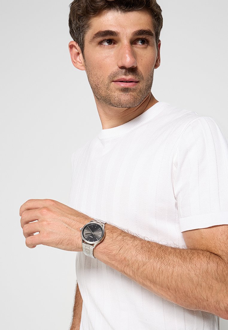 Montre en argent avec un cadran noir, boîtier rond et un bracelet métallique. Le modèle porte une chemise blanche à manches courtes et texturée. Arrière-plan neutre.