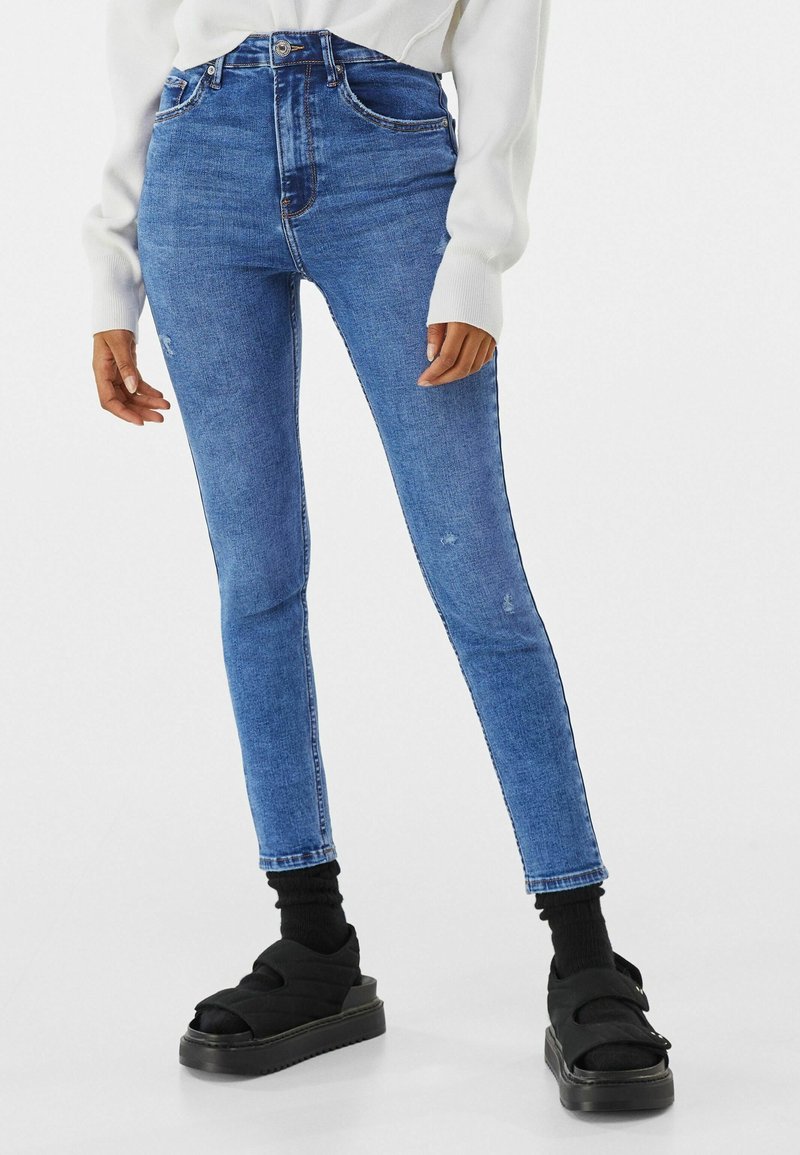 Person trägt enge blaue Jeans, schwarze Socken, schwarze Plateausandalen und ein weißes langärmeliges Oberteil und steht vor einem einfarbigen Hintergrund.