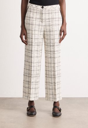 Femme portant un pantalon blanc taille haute à jambes larges avec un motif à carreaux noirs, un bouton noir et des chaussures Mary Jane noires avec des boucles dorées.