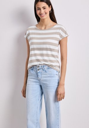 Kvinde smilende iført beige og hvidstribet kortærmet skjorte, stoppet ned i lyseblå højtaljede jeans, stående mod en ensfarvet væg.