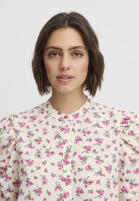 Kremowa bluzka w kwiaty w różowe wzory, z bufiastymi rękawami i okrągłym dekoltem. Posiada różowe guziki na przodzie.