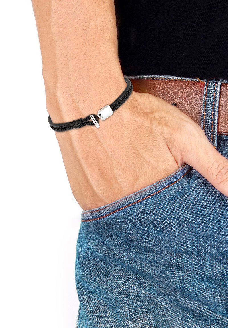 Brățară împletită neagră cu un închizător elegant din argint, așezată pe o încheietură, asociată cu blugi albastri și o curea de piele maro. Textură netedă, design minimalist.