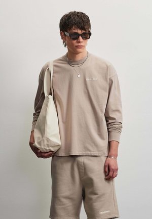 Young man wearing beige long-sleeve shirt and shorts with text, black sunglasses, silver necklaces, and carrying a white tote bag on shoulder.