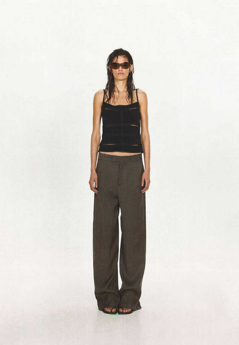 Femme aux cheveux mouillés portant des lunettes de soleil foncées, un haut noir sans manches, un pantalon gris ample et des sandales noires, debout devant un fond clair uni.