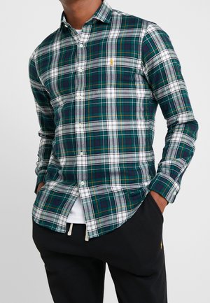 Homme portant une chemise à carreaux vert, marine, blanc et jaune avec un pantalon noir, les mains dans les poches, sur un fond uni.