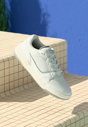Vita lädersneakers med snören som vilar på gula kakeltrappor, med en livlig blå vägg i bakgrunden i starkt dagsljus.